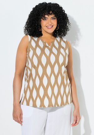 Frau mit lockigem schwarzen Haar, die ein ärmelloses beiges und weiß gemustertes Oberteil sowie weiße Hosen trägt, lächelt vor weißem Hintergrund.