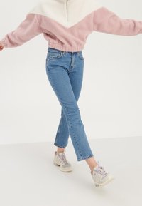 Rosa-weißer Fleece-Pullover mit halbhohem Reißverschluss, kombiniert mit blauen Jeans mit geradem Bein und hellgrauen Turnschuhen mit lila Akzenten.