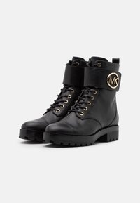Botas de tobillo de cuero negro con puntera redonda, acentos de hardware dorado, cordones en la parte delantera y una gran correa con un logotipo circular. Suela gruesa y resistente.
