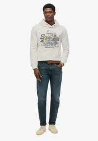 Sudadera gris claro con estampado gráfico, vaqueros azules y zapatillas beiges. La sudadera cuenta con un cordón y un bolsillo tipo canguro. Paleta de colores neutros.