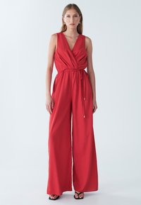 Rød ærmeløs heldragt med dyb V-udskæring, bindebånd i taljen og bredt ben, lavet af letvægtsstof. Glat tekstur og minimal detaljering.