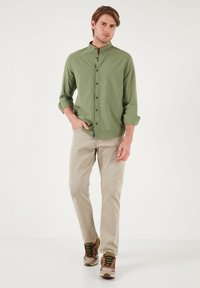 Chemise verte à manches longues avec col mao, fermeture à boutons sur le devant, pantalon beige et baskets à motif camouflage, se tenant sur un fond blanc.