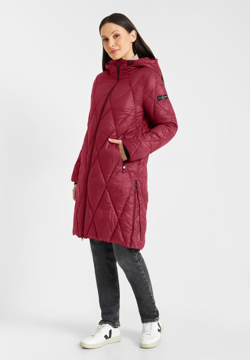 Vrouw staat in een lange rode gewatteerde jas met capuchon, zwarte jeans en witte sneakers, met één hand in haar zak.