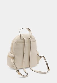 Beige Leder-Rucksack in runder Form, mit zwei Schulterriemen, einem oberen Griff und goldfarbenen Reißverschlüssen. Verfügt über seitliche Reißverschlusstaschen.
