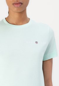 Mėtų spalvos medvilninis marškinėliai su apvalia iškirpte ir trumpomis rankovėmis, su balta "GANT 1948" logo ir mažą šarvų akcentą krūtinėje.