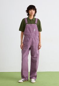 Salopette larghi color lavanda con spalline regolabili, tasche frontali e un patch con logo, abbinati a una polo verde a trama e sneakers bianche.
