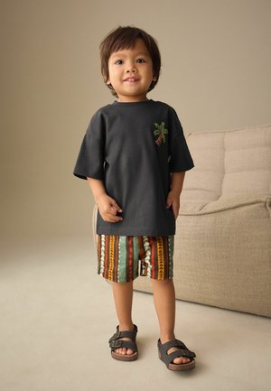 Kleines Kind steht neben einem beigen Sofa, trägt ein schwarzes T-Shirt mit Palmenmotiv, gemusterte Shorts und schwarze Sandalen mit Schnalle.