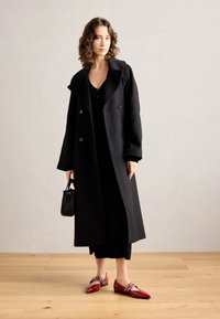 Schwarzer doppelt genähter Mantel mit breiten Revers, getragen über einem dunklen Outfit. Glänzende rote spitze Flats mit Akzentriemen. Kleine schwarze Handtasche.