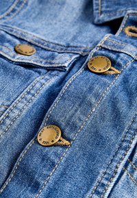 Detailní záběr na modrou džínovou bundu s viditelnými bronzovými knoflíky a žlutým prošíváním na švech a klopě kapsy.