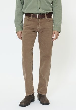 Mand iført brune kordfløjlsbukser, mørkebrune støvler og en grøn langærmet skjorte med et brunt bælte, stående mod en hvid baggrund.