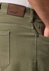 Pantalones verde oliva hechos de una mezcla de algodón con una etiqueta de cuero marrón que dice "MEN+". Las características incluyen costuras y bolsillos laterales.