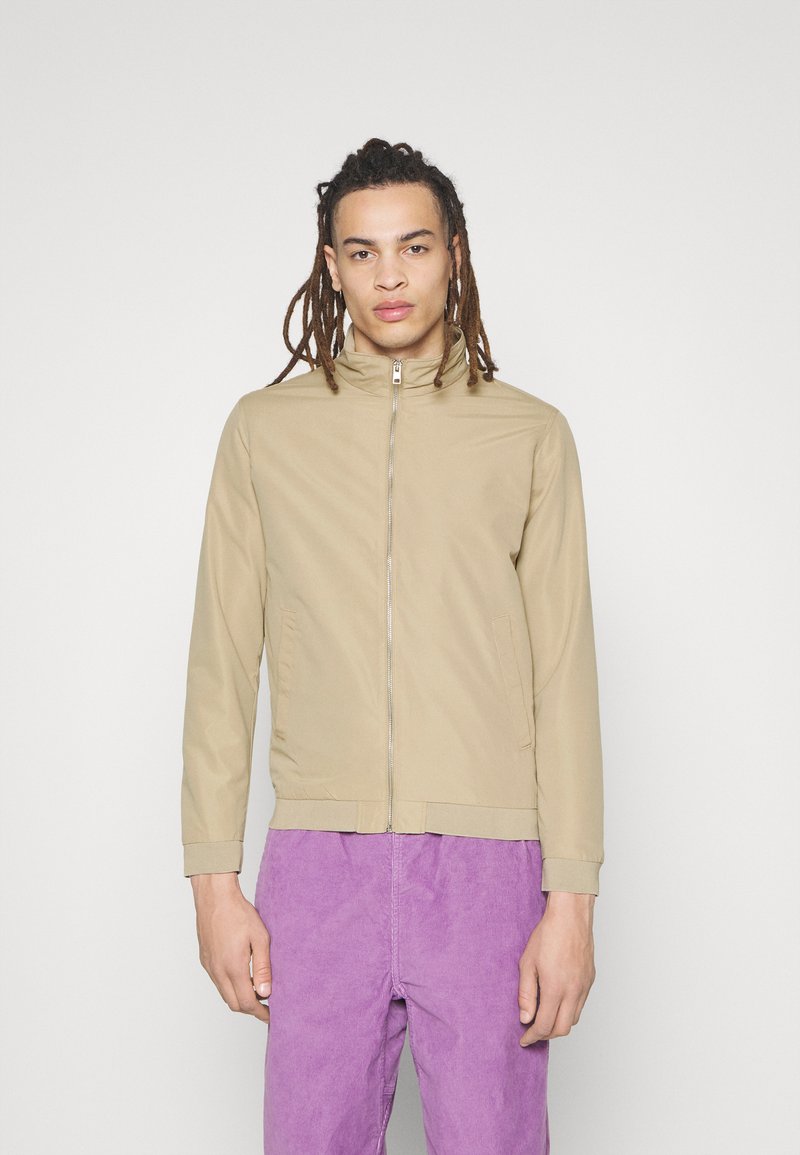 Giacca beige chiaro con colletto alto, zip frontale e tasche laterali. Tessuto liscio, vestibilità affusolata, abbinata a pantaloni in velluto a coste viola.