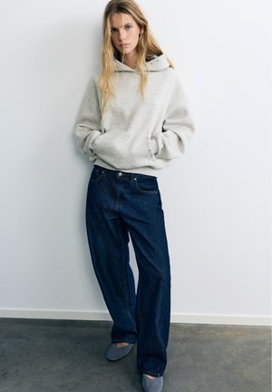 Jeune femme aux longs cheveux blonds portant un sweat à capuche gris clair, un jean large bleu foncé et des chaussures plates bleues, debout contre un mur blanc.