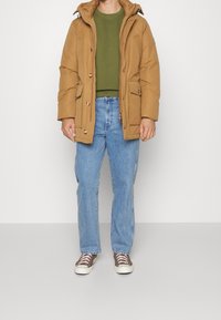 Giacca imbottita marrone con cappuccio, con chiusura frontale con cerniera e bottoni, indossata sopra una felpa verde e jeans blu. Scarpe da ginnastica marroni.