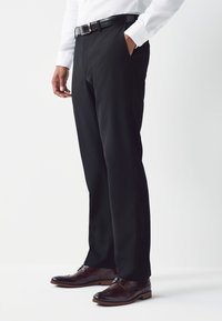 Pantalon noir sur mesure à la texture lisse, associé à des chaussures brogues marron. Une ceinture en cuir noir souligne la taille.