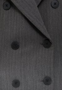 Manteau croisé gris en chevron avec boutons noirs et revers crantés, présentant un tissu texturé et une coupe ajustée.