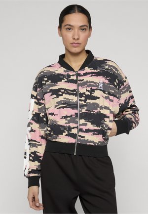 Kvinne iført en bomberjakke med digitalt kamuflasjemønster i beige, rosa og svart, med svart krage og mansjetter, stående med den ene hånden i lommen.