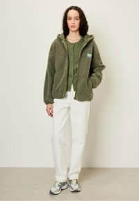 Veste en polaire vert olive doublée, avec capuche, fermeture éclair et patch logo, superposée à un pull vert à boutons, accompagnée de jeans blancs et de baskets.