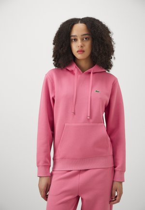 Rosa Kapuzenpullover aus weichem Stoff, mit einer Fronttasche, Kordeln und einem kleinen grünen Logo. Kombiniert mit passenden rosa Hosen.