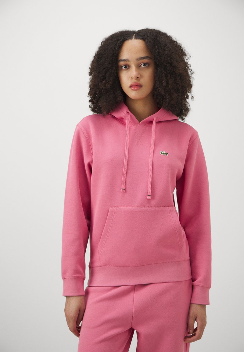 Felpa rosa realizzata in tessuto morbido, con tasca frontale, coulisse e un piccolo logo verde. Abbinata a pantaloni rosa coordinati.