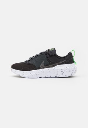 Chaussure de sport noire avec lacets gris, languette de traction verte, swoosh gris à motifs et semelle épaisse blanche à points.