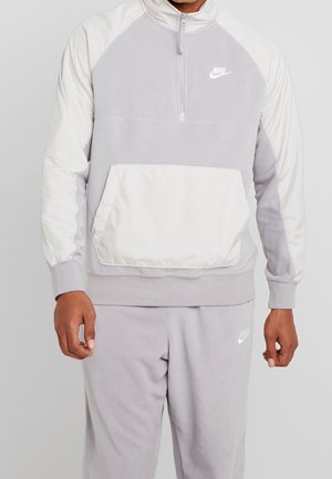 Mężczyzna ubrany w jasnoszary polar Nike z białym panelem i dopasowane spodnie dresowe, stojący na jednolitym tle.