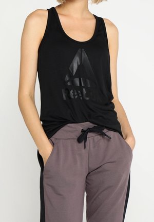 Kvinde iført sort, ærmeløs Reebok tanktop og grå bukser med snøre, med hænderne i lommerne, foran hvid baggrund.