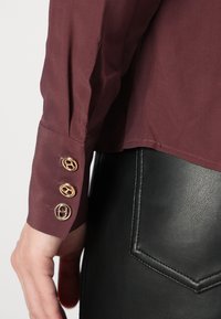 Maroon blouse with three gold button accents at the cuff. Smooth fabric with a subtle sheen, paired with a black textured leather garment.