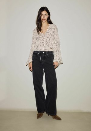 Woman with long dark hair wearing a beige polka-dot blouse, high-waisted black wide-leg jeans, and brown pointed boots stands against a plain wall.
