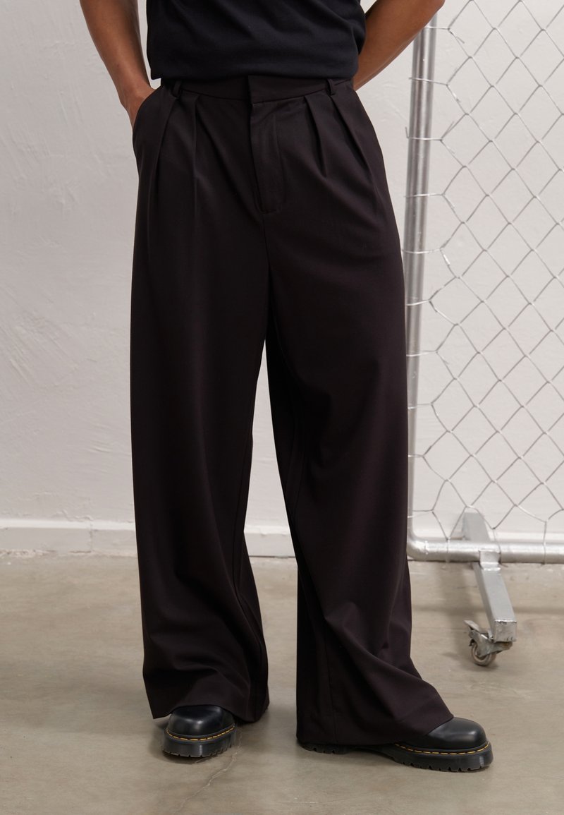 Personne portant un pantalon noir à jambes larges avec des plis latéraux et des chaussures noires à semelles épaisses, debout les mains dans les poches près d'une clôture métallique.
