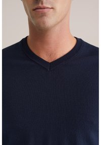 Tmavě modrý svetr s výstřihem do véčka, vyrobený z měkké pletené látky. Výrazné detaily zahrnují žebrovaný výstřih a hladkou texturu.