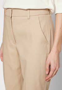 Beżowe spodni z materiału o gładkiej fakturze, z widocznymi kieszeniami z przodu i płaskim pasem. Tkanina wydaje się lekka i przewiewna.