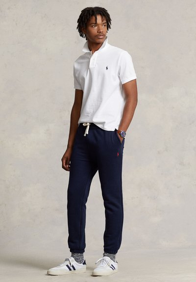 Jaunas vyras stovi, dėvi baltą polo marškinėlį, tamsiai mėlynas joggerius su raišteliu, raštuotas kojines, baltus sportinius batelius ir laikrodį ant riešo, neutrali fonas.