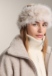 Világos bézs műszőrme fejpánt, bolyhos textúrával. Krémszínű bordázott garbóval és lágy bézs műszőrmével bélelt dzsekivel viselve.