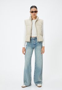 Beige gepolsterte ärmellose Weste, weißes Langarmshirt, hellblaue weit geschnittene Jeans und weiße Sneakers mit braunen Sohlen.