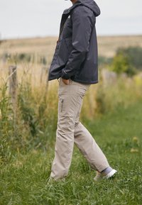Person in beige cargo pants and dark jacket walking on grassy path near wooden fence in a natural outdoor setting.