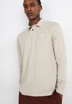 Beige long-sleeve polo shirt with a collar, featuring a small logo patch and two black buttons at the neck. Smooth, lightweight fabric.