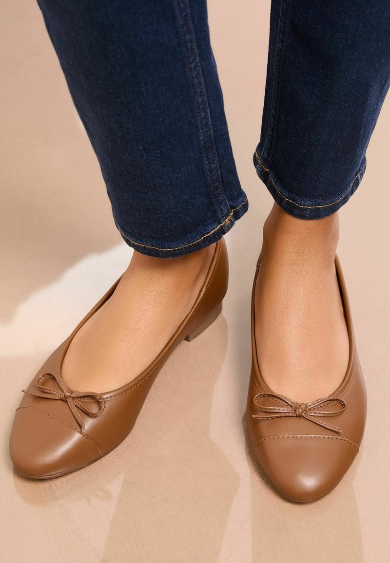 Vrouw draagt bruine leren platte schoenen met kleine strikjes, gecombineerd met donkerblauwe spijkerbroek, staand op een beige vloer.