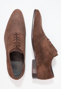 Chaussures en suede marron avec un bout pointu, six œillets et une semelle intérieure en cuir lisse. Elles présentent un talon bas en bois foncé et une finition texturée solide.