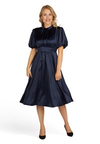 Marineblauwe satijnen jurk met pofmouwen en een geraffineerde taille, met een rok tot op de knie. De gladde textuur weerkaatst het licht subtiel.