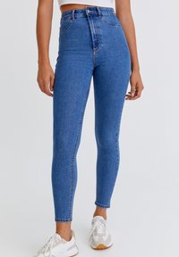 Jean en denim bleu taille haute, coupe slim, avec un design classique à cinq poches et un délavage subtil, assorti à des baskets blanches sportives.