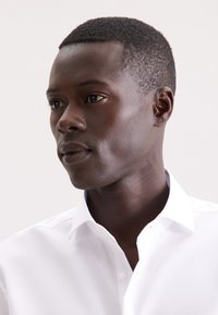 Camicetta bianca con colletto, texture liscia e fresca; presenta un collo con bottoni e una vestibilità su misura. La tonalità della pelle appare scura.