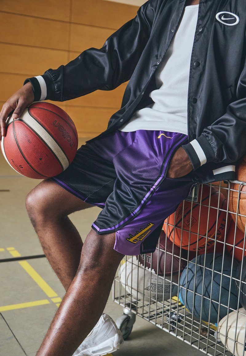 Personne en veste noire et short de basket violet tenant un ballon, assise sur un chariot rempli de divers ballons de sport dans une salle de sport.