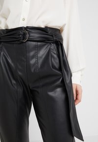 Zwarte faux leren broek met een geknoopte riem met een ronde gesp en plooien, aangevuld met een witte overhemden.