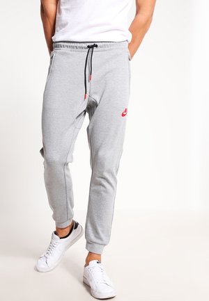 Mężczyzna ubrany w jasnoszare spodnie dresowe Nike z czarnym sznurkiem, białe sneakersy Nike oraz białą koszulkę stojący na jednolitym tle.