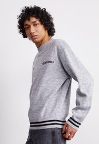 Grå sweatshirt med ribbet rundhals og sorte stribede manchetter. Har broderet mærke-logo i sort på forsiden. Materialet er en bomuldsblanding.