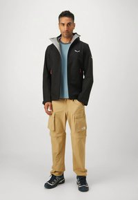 Juoda neperšlampama striukė su pilku pamušalu, oranžiniais užtrauktukų traukikliais; derinama su rudos spalvos krovinio kelnėmis su didelėmis šoninėmis kišenėmis ir mėlynomis žygių batais.