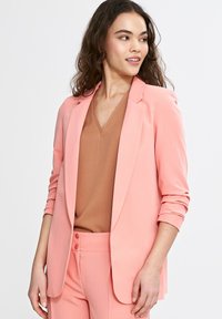Jasnoróżowa marynarka o dopasowanym fasonie, z klapami w kształcie litery notched i podwiniętymi rękawami. Nosi się ją na brązowej bluzce z dekoltem w serek. Gładki materiał z dopasowanym krojem.