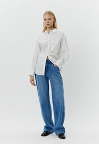 Bijela pamučna košulja na dugmad s džepom, uparena s širokim plavim trapericama i crnim cipelama. Glatka tekstura, opušteni stil.