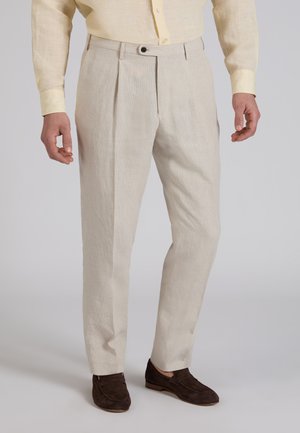 Hombre con pantalones beige plisados, camisa amarilla pálida abotonada y mocasines de ante marrón oscuro, sobre un fondo liso.